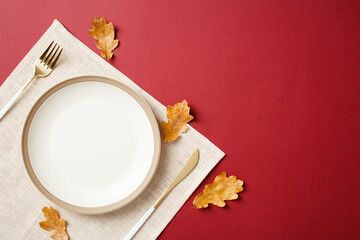 Elegant autumn table setting with white empty plate, gold cutlery, and scattered fall leaves on red...