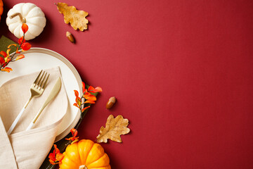 Autumn table setting with white plate, gold cutlery, beige napkin, and decorations of pumpkins, acorns, and fall branches on a deep red background. Thanksgiving dinner concept.