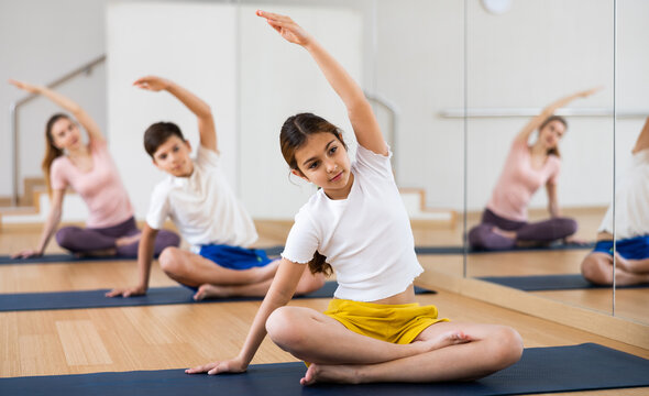 Smiling teen girl doing Padmasana with side stretch during yoga workout in gym with family..