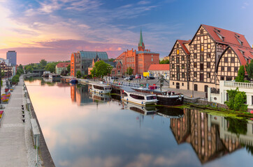 Panoramic view of the Bydgoszcz waterfront at sunrise with boats along the Brda River, historic half-timbered buildings, and modern structures in Poland