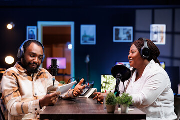 African American podcast host interviews a female content creator in a neon-lit home studio using advanced recording equipment. Their engaging discussion is broadcast digitally to their online fans.
