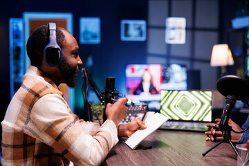 Fototapeta premium Male talk show host holding a notebook and interviewing a guest, having online discussion for their listeners. African american man using recording equipment to create a radio program at home.
