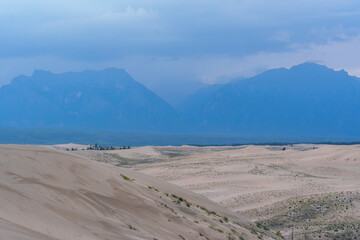 Expansive desert dunes under a dramatic sky with distant mountains