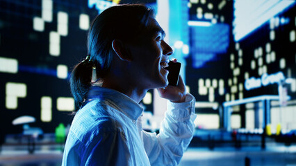 Asian man wandering around city avenues during nighttime, talking over the phone with friend while looking upwards to surrounding buildings in metropolitan urban environment