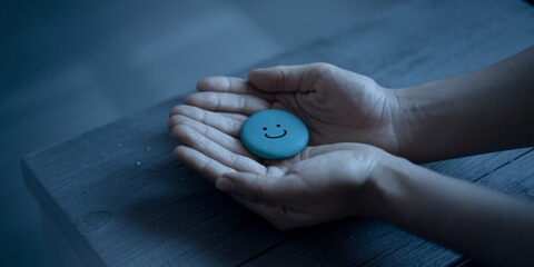 Fototapeta premium A photo of two hands gently cradling a teal circle with a simple smiling face drawn on it. The hands are placed on a wooden surface. 