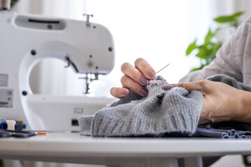 A woman uses her hands to sew up a hole in a gray knitted garment with a needle and thread. 