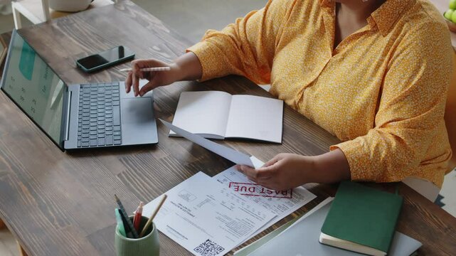 Close-up high angle view of unrecognisable Black woman checking deadlines for payment of utility bills using laptop and writing data down on sheet of paper sitting at desk