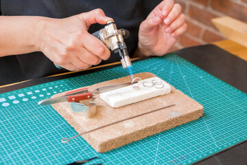 A woman uses a blowtorch to create jewelry on a on a worktable with cutting mat.