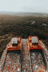 Two orange recycling bins filled with plastic bottles and other recyclable materials, overlooking a forest and city skyline.