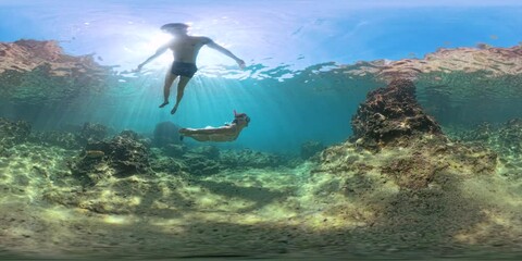 Panorama spherical VR. Two people swim underwater in clear blue water, exploring coral and rocks. Sunlight filters through the surface in a 360 view, creating a stunning immersive scene.