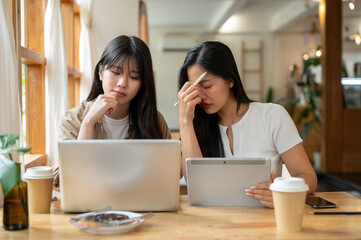 Obraz premium Two serious and stressed Asian female friends are working remotely together from a coffee shop.