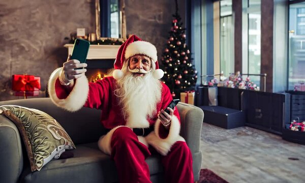 P&egrave;re No&euml;l assis sur une banquette dans un salon de style industriel, prenant des selfies ou faisant une visioconf&eacute;rence sur deux portables, image d'un P&egrave;re No&euml;l accro aux &eacute;crans