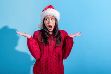Obraz premium An Asian woman wearing a red knit sweater and a Santa hat stands with her hands raised in a surprised. Her mouth is open in an expressive. The background is light blue.