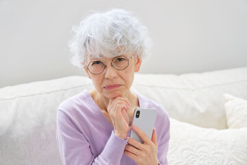 スマートフォンを持って悩むシニア女性　リビングルーム　カメラ目線