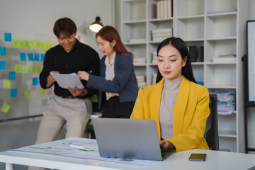 Focused on the Task: A young woman in a vibrant yellow blazer works diligently on her laptop, while her colleagues discuss plans behind her.
