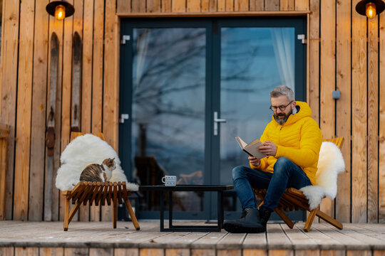 Mature man sitting on terrace reading book, enjoying cup of coffee during cold winter day. Peaceful moment alone in cabin.