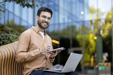 Young man smiling using laptop outdoors. Represents remote work, technology, productivity, and...