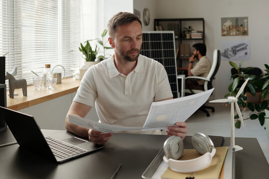 Serious businessman sitting by workplace in coworking space and looking through documents with project materials