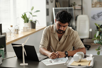 Bearded engineer sitting by workplace with laptop and looking through text on paper with windmill turbine