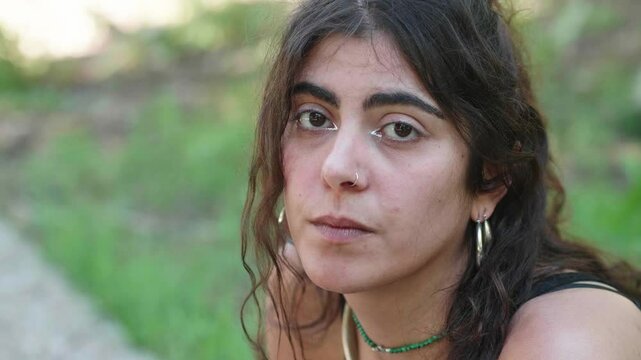 A brunette woman with straight hair wearing a black shirt sits discontentedly in the park, feeling unfulfilled and disappointed with life, capturing a moment of deep reflection.
