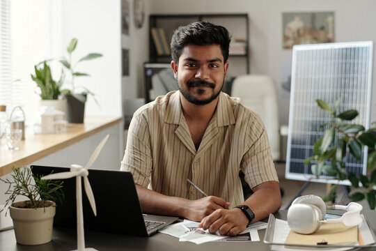 Happy young male architect looking at camera while sitting by workplace and drawing sketch for new project