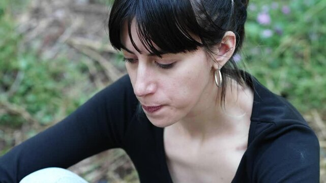 A brunette woman with straight hair wearing a black shirt sits with her knees up in the park, looking dejected and disheartened by her colleagues' behavior, capturing a moment of sorrow.