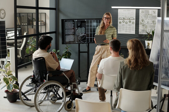 Young female coach standing by interactive board and communicating with group of intercultural colleagues