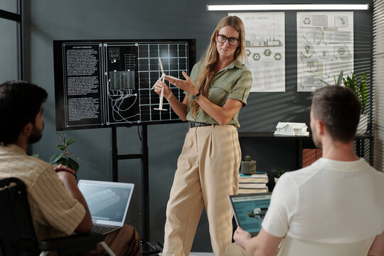 Young female coach with model of windmill standing by interactive board and making presentation to male colleagues
