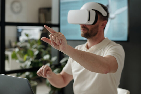 Focus on hand of modern businessman wearing VR helmet pointing at virtual screen or mockup during presentation