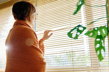 Mature woman wrapped in blanket looking through window blinders at home
