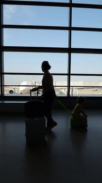 Silhouetted view, woman walks through airport arrival passage, pulling her toddler son on ride-on suitcase. Slow motion side tracking shot, post-flight time as they head towards immigration