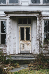Front entrance of an abandoned, weathered house with broken windows and overgrown vegetation, radiating a spooky and desolate atmosphere.