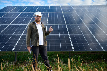 Engineer near solar station showing thumb up gesture. Supervisor is checking on solar panels on solar farm. Solar and renewable energy. Front view.