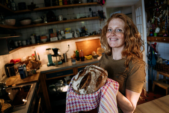 Smiling woman holding baked bread in kitchen at home