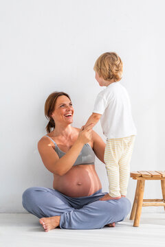 Blond boy standing on pregnant mother's lap near white wall