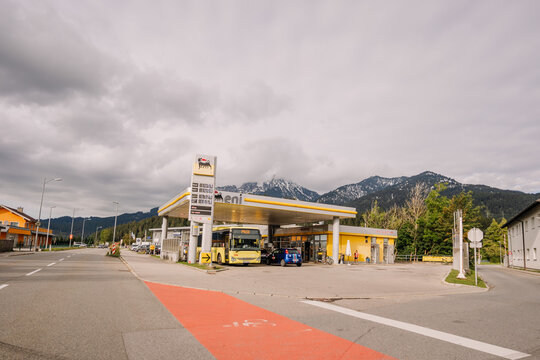 A gas station with a bus and cars parked under the roof, with mountains and cloudy skies in the background..