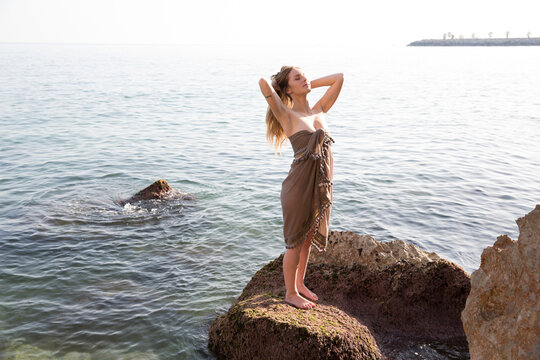 Young woman wearing sarong and standing with hand in hair on rock