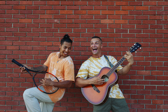 Cheerful brother enjoying leisure time with guitar and tennis racket in front of brick wall
