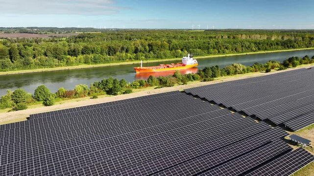 Tanker transporting fossil fuels along  waterway, sailing past canal bordered by solar farm. Traditional and modern energy. Concept of  transition from fossil fuels to renewable energy.