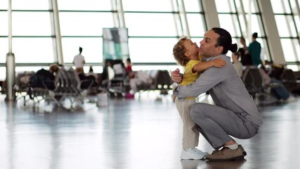 Little boy come with open arms and hugs his father, who is crouched down, welcoming him with arms wide open too. Touching scene of embrace, in boarding area of airport, side view, blurred background