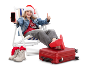 Christmas Holiday travel and vacation concept, tourist woman in travel attire wears a santa hat, on deck chair with trolley suitcases, showing smartphone and thumbs-up. Flight and travel booking.