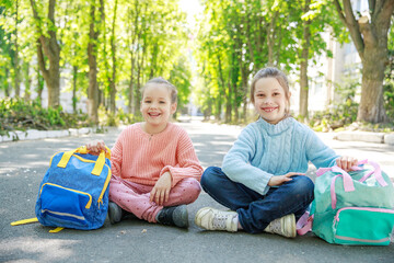 Two joyful girls sit cross-legged on a quiet street, basking in the sunlight. The vibrant greenery around them enhances their playful spirit, making for a delightful afternoon.