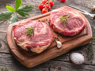 Raw beef steaks with herbs and spices ready for cooking are on wooden board.
