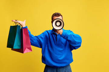 Stylish man lifting colorful bags with megaphone in hand, announcing shopping deals against sunny yellow background. Concept of discount deals, shopping spree, Black Friday, marketing promotion.