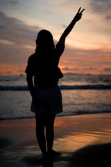 Fototapeta premium Silhouette of girl posed raising peace hand. Silhouette of woman on the beach at sunset