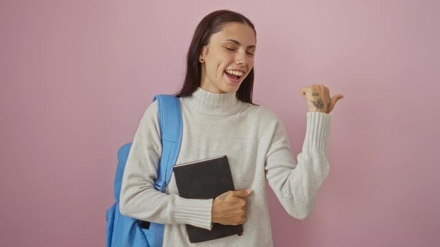 Student, young hispanic woman holding book and backpack, pointing to the side with thumb up and smiling happily over isolated pink background with open mouth