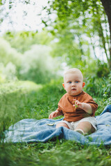 A blond toddler in a t-shirt and shorts sits on the green grass and plays. A child is playing on the street. The baby sits independently.