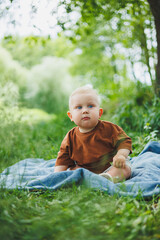 A blond toddler in a t-shirt and shorts sits on the green grass and plays. A child is playing on the street. The baby sits independently.