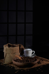 Photo of a coffee grinder with a bag of coffee beans, a coffee spoon, a cup of coffee and coffee beans on a dark window background