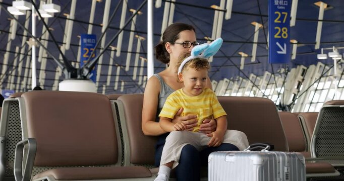 Cute toddler boy with bunny ears headband sits on his mother's lap in airport, waiting near boarding gate. Slow-motion portrait captures rare calm moment of usually playful son.